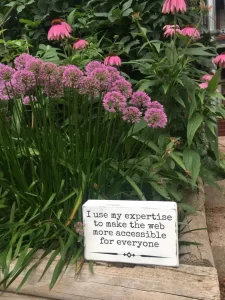 A wooden sign rests in a flower bed and reads "I use my expertise to make the web more accessible to everyone."